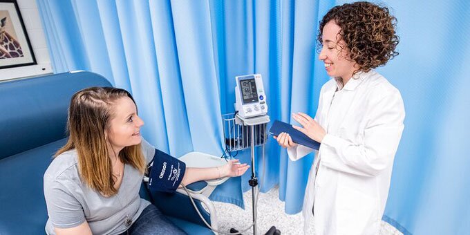 Two students speaking, one is getting their blood pressure taken while the other has a lab coat on