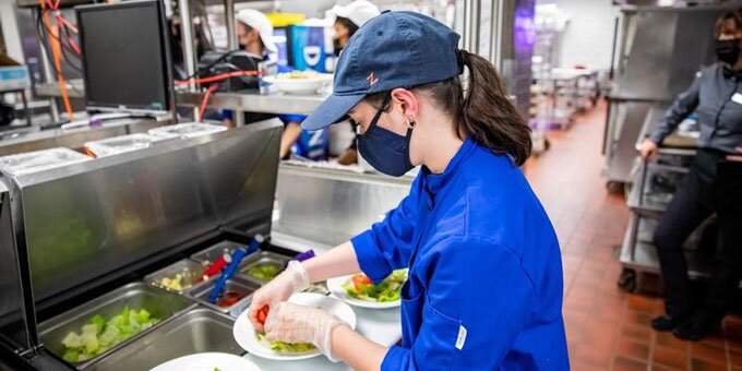 Student making a salad in a kitchen with a blue lab coat on